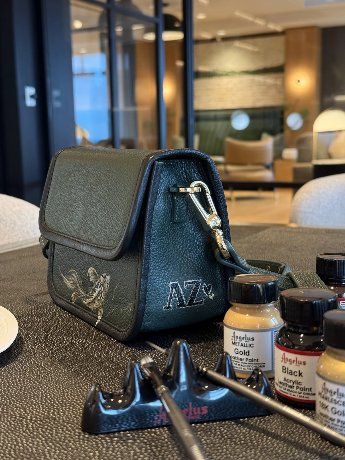 A painted koi on the front of a dark green leather bag, with AZ initials on its side, beside Annem’s paints.